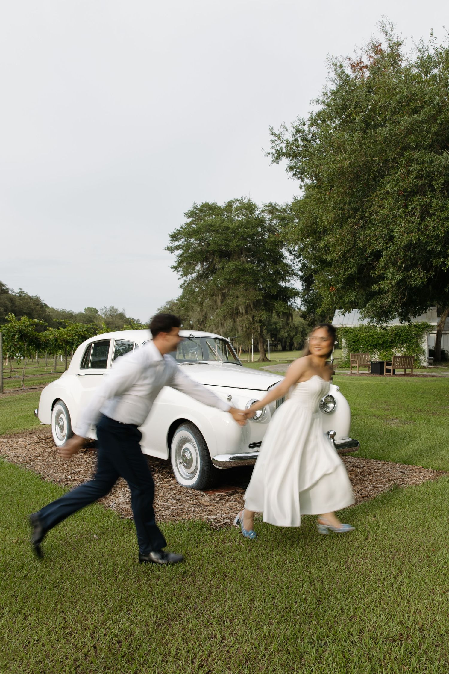Bride and groom running hand-in-hand past a classic white vintage car on a green lawn at an outdoor wedding venue.