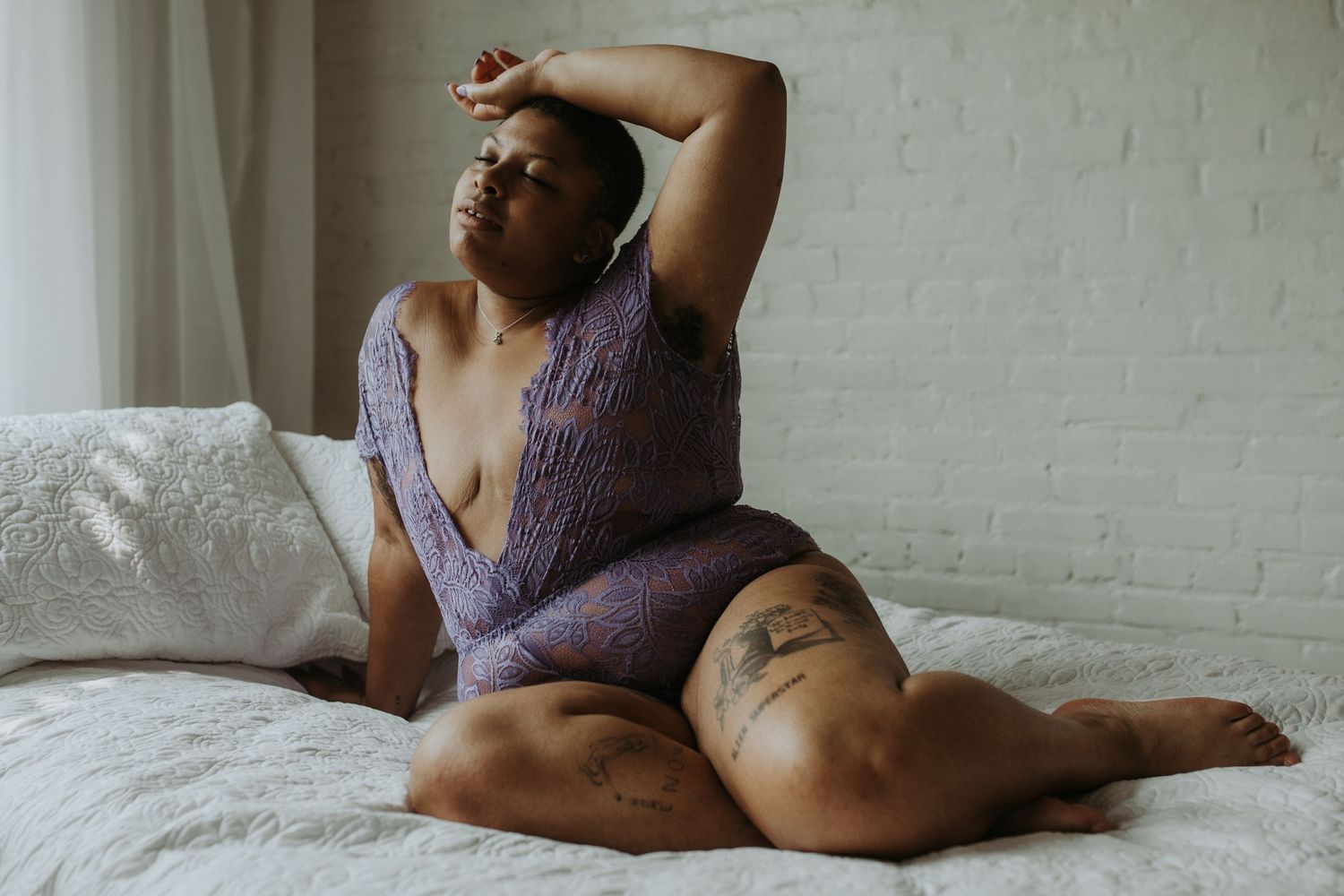 A plus-size woman in a purple lace bodysuit poses gracefully on a white bed in a bright room.