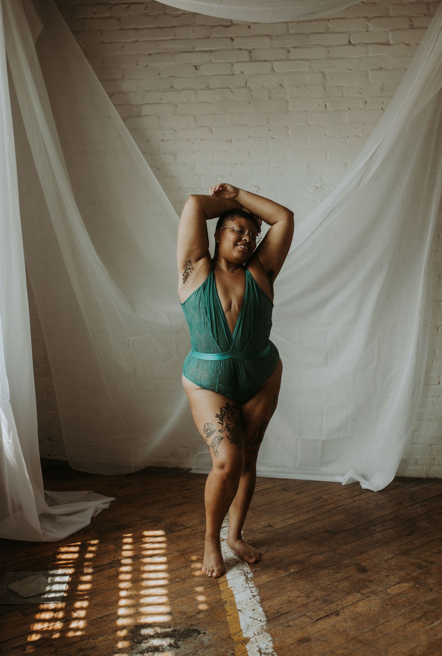 A plus-size woman poses confidently in a teal green lingerie bodysuit with tattoos, standing before white sheer curtains.