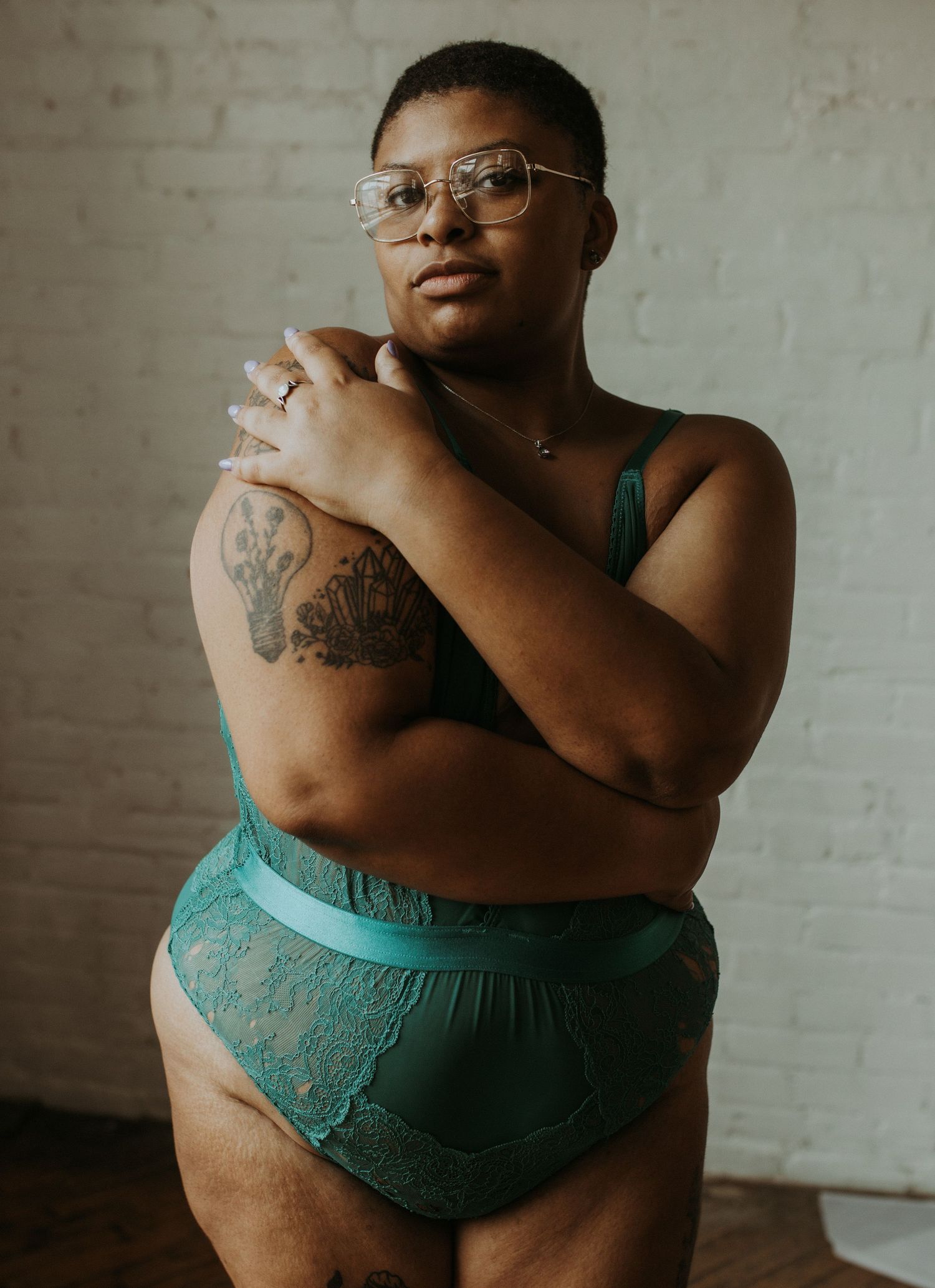 Plus-size model in teal green lace lingerie set, wearing glasses, with arm tattoo, posing confidently indoors.