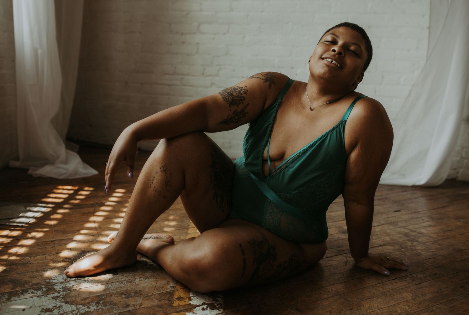 A plus-size woman in a teal bodysuit poses confidently on a wooden floor in natural light.