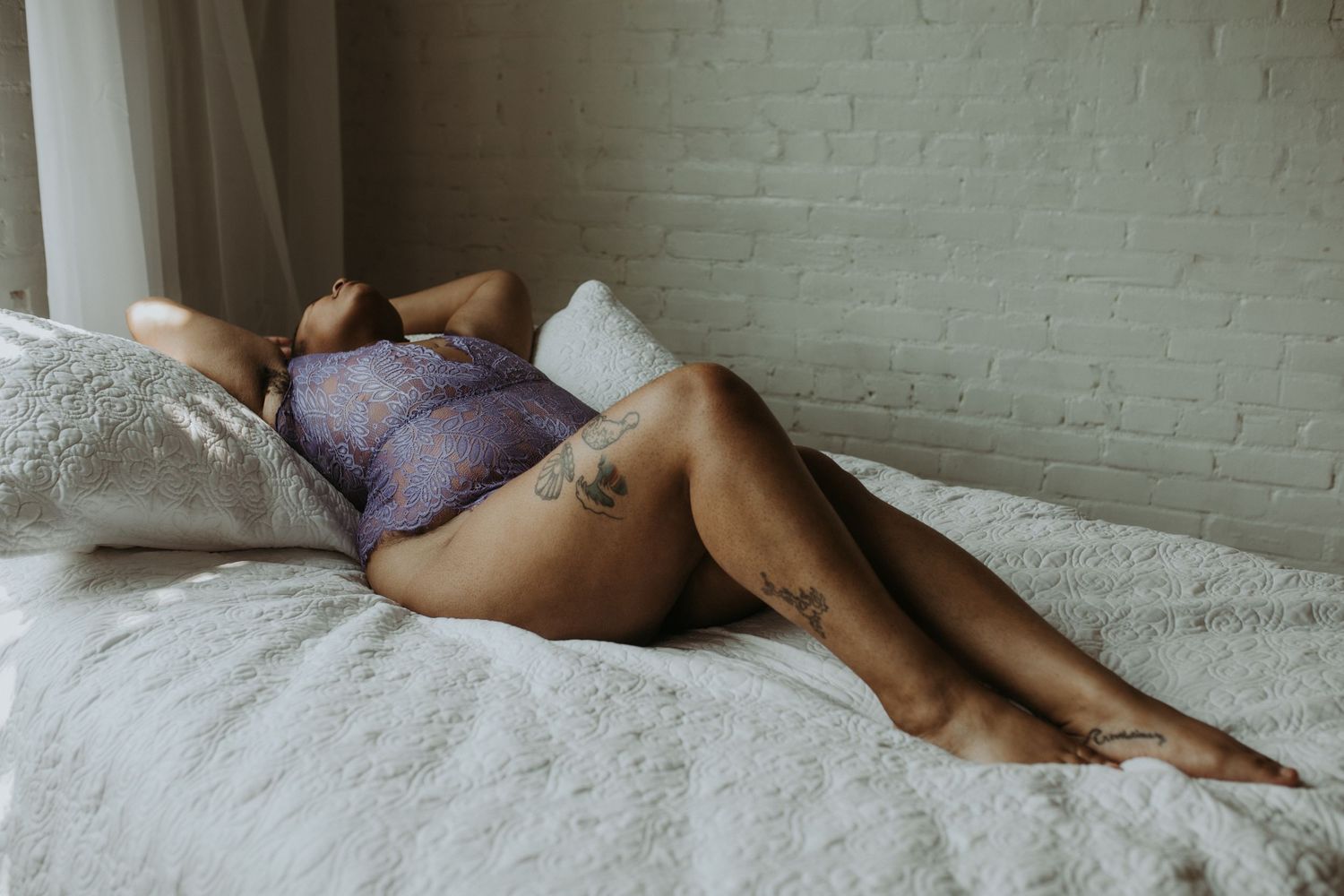 A woman in purple lace lingerie reclines gracefully on a white bed, showcasing tattoos in a boudoir setting.