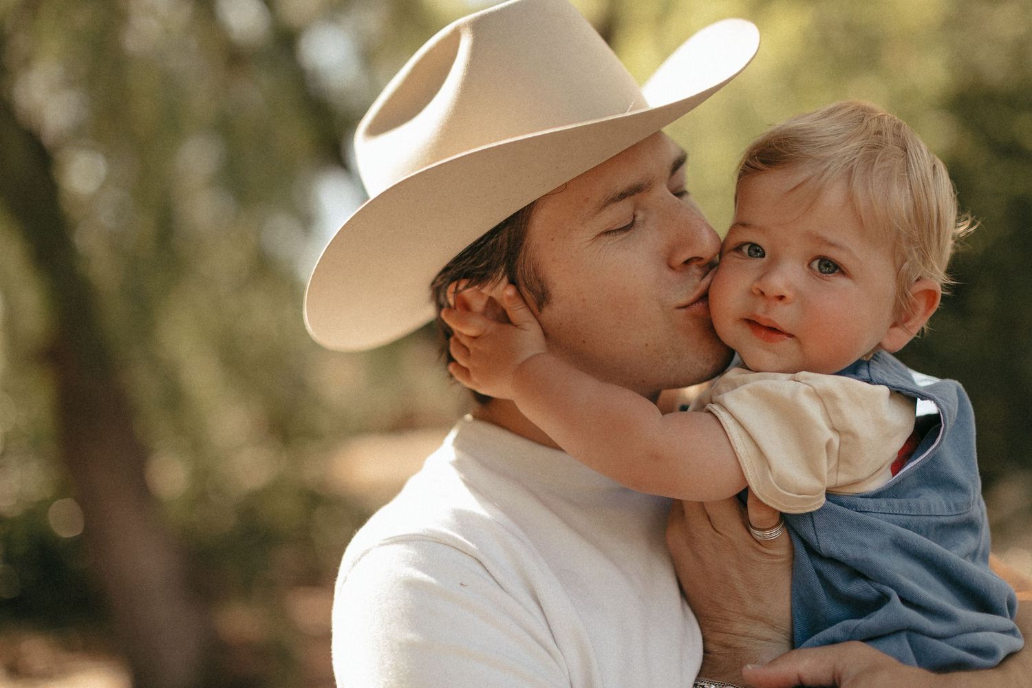 A man in a white cowboy hat kisses a cute toddler boy outdoors with green trees in the background.