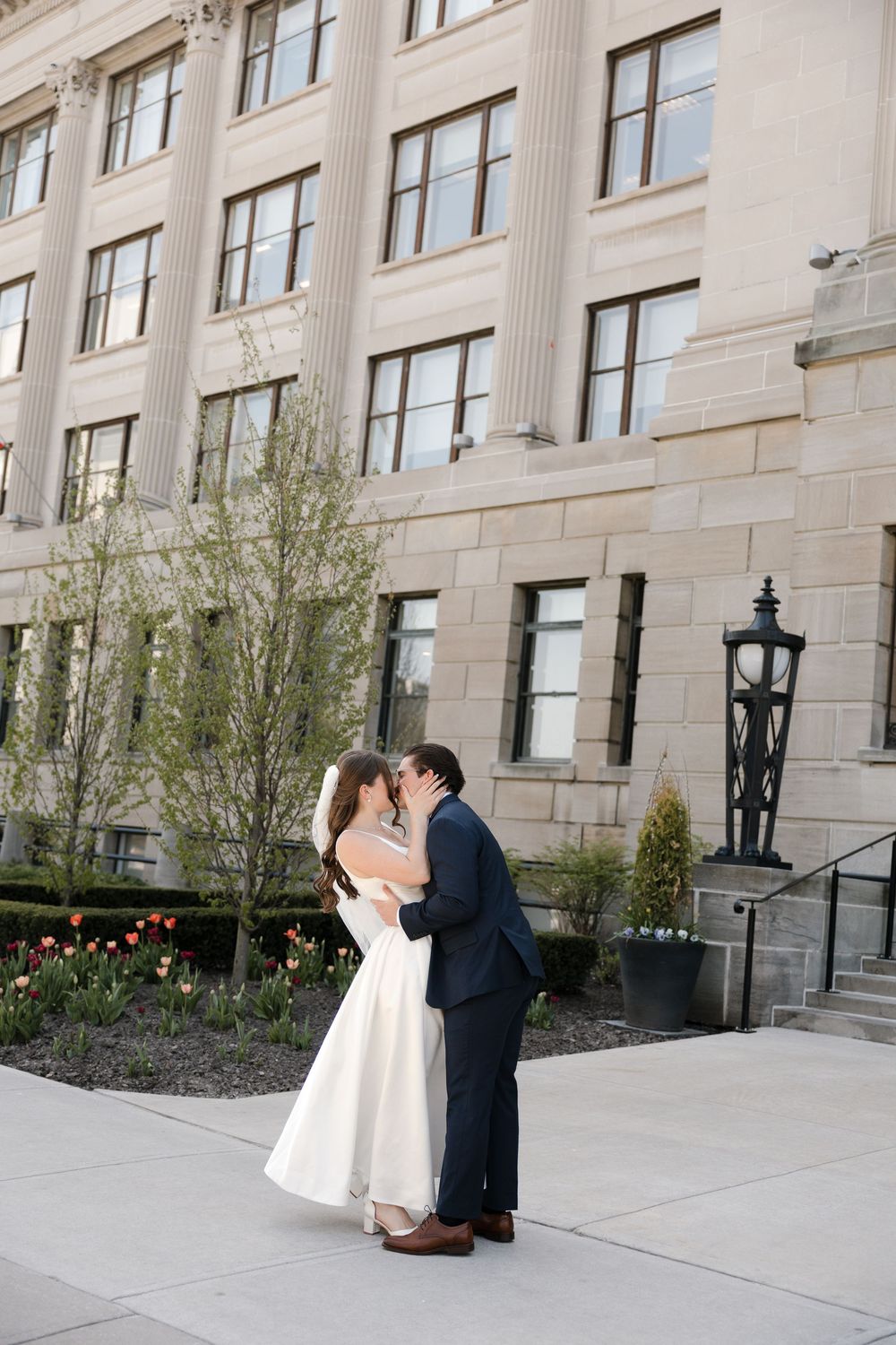 Bride and groom share romantic kiss outside elegant stone building with spring tulips in bloom.