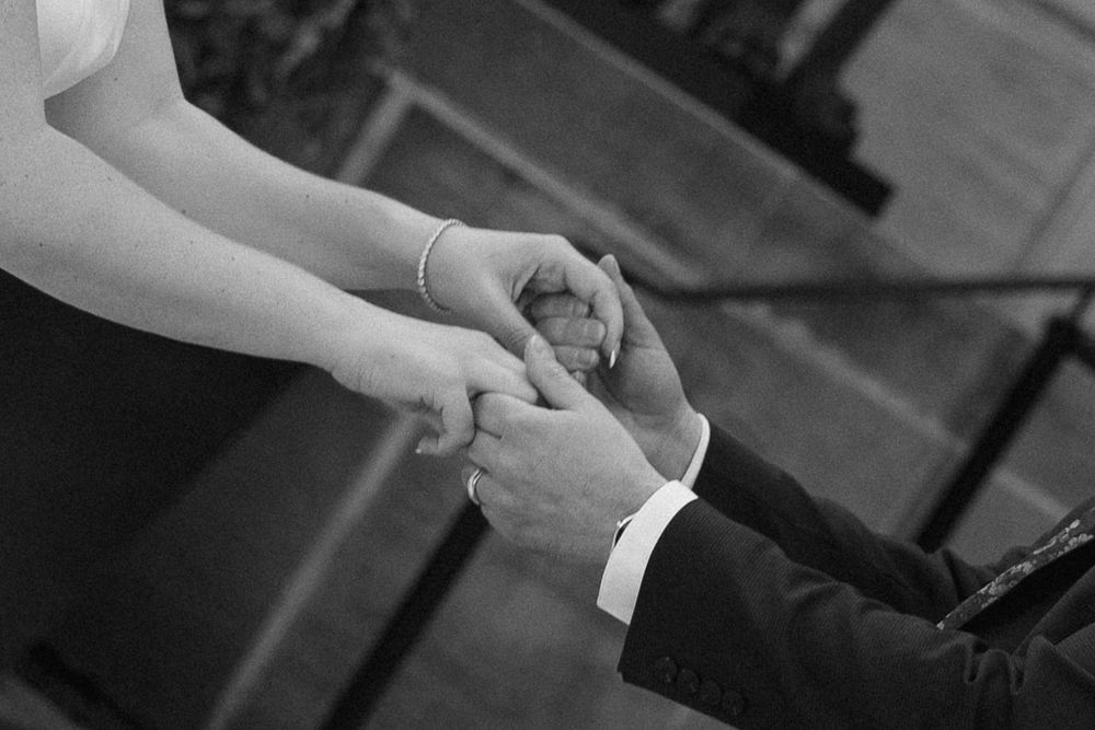 Black and white close-up of bride and groom gently holding hands during their wedding ceremony.