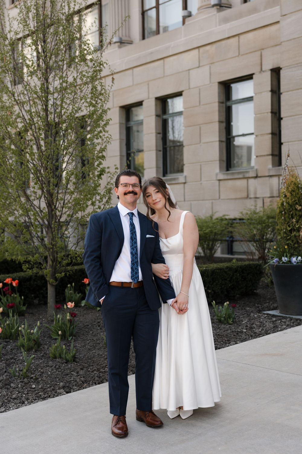 Bride and groom pose together outdoors in front of a stone building surrounded by spring tulips.