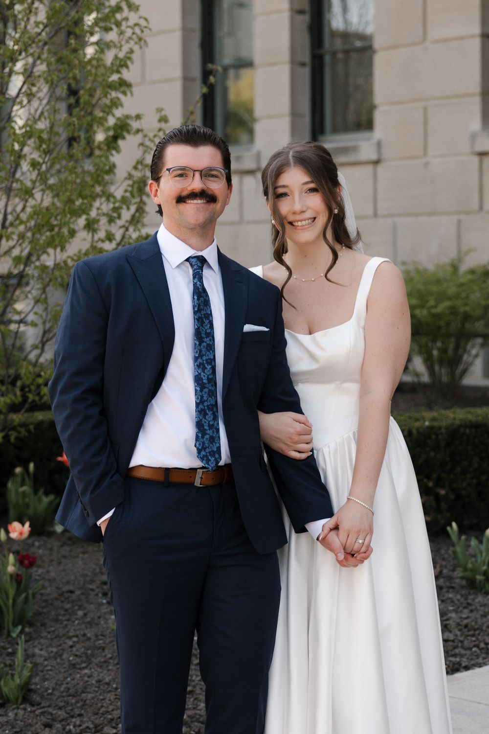 Smiling bride and groom with mustache pose together outdoors near a stone building with spring flowers.