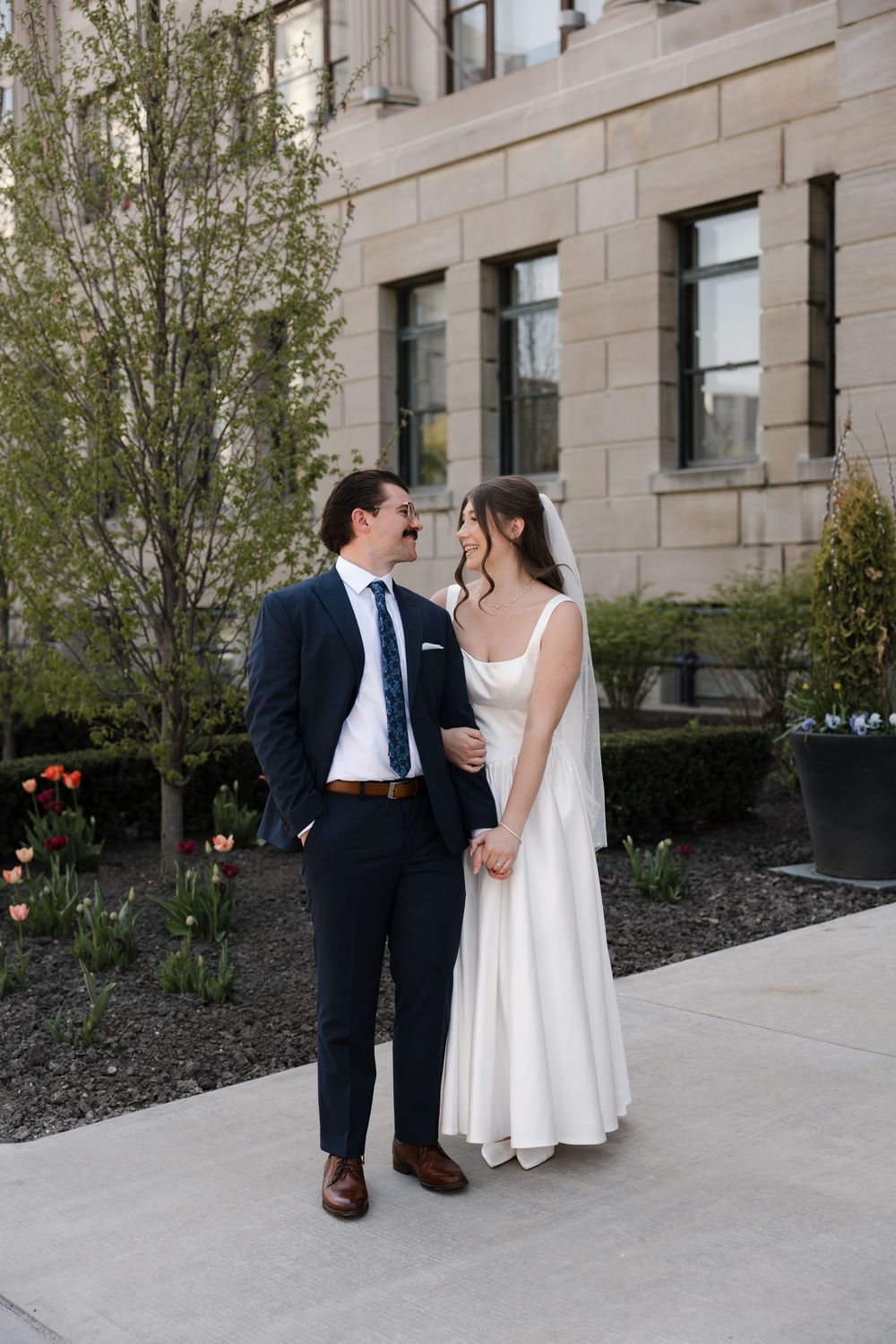 Bride and groom walking hand in hand past tulips outside stone building.