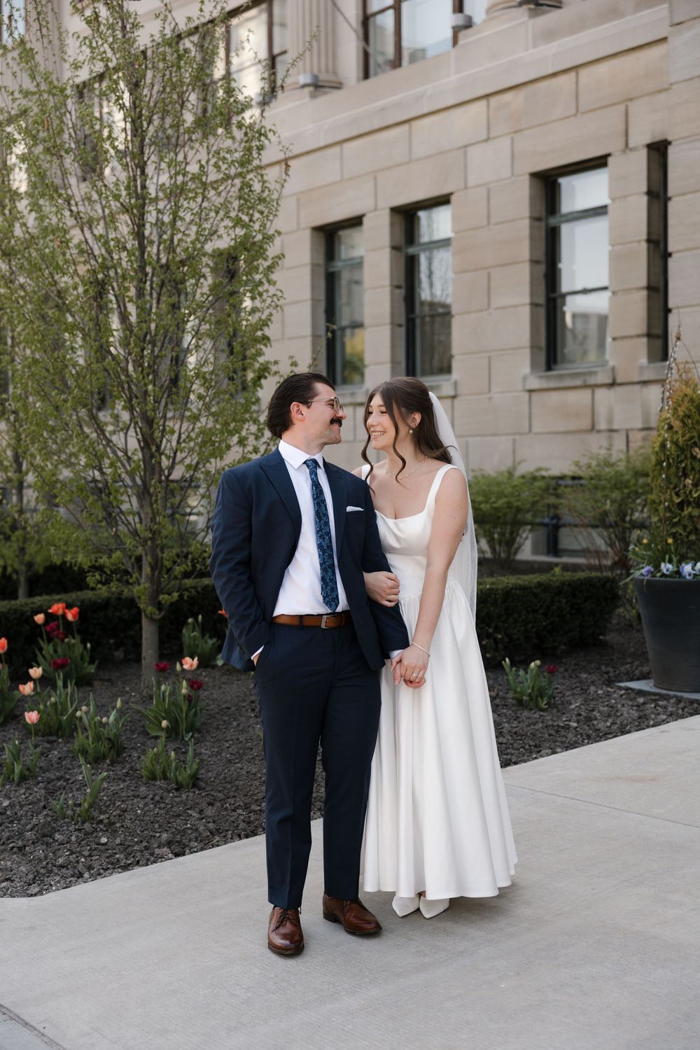 Bride in white dress and groom in navy suit holding hands outside historic stone building.