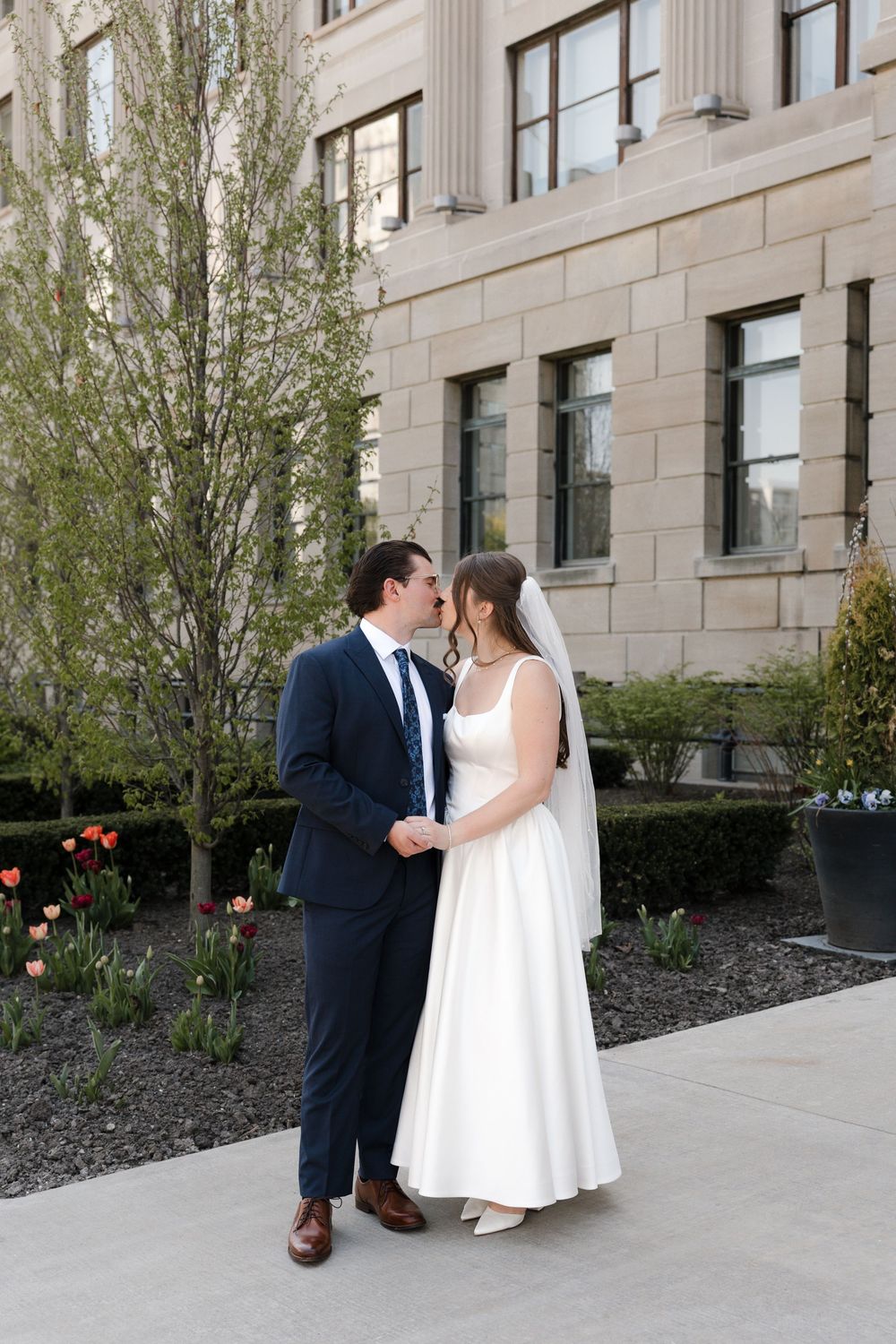 Bride in white gown and veiled groom in navy suit share a romantic moment outside a stone building.