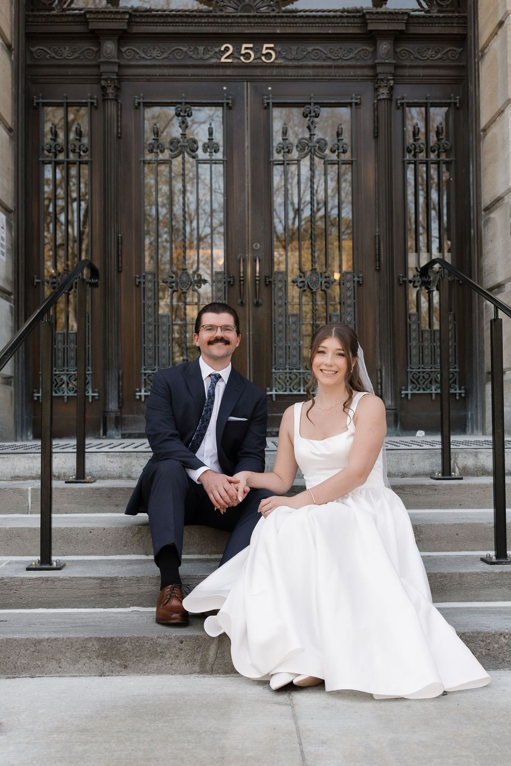Bride and groom sitting on steps of an ornate building entrance numbered 255 in wedding attire.