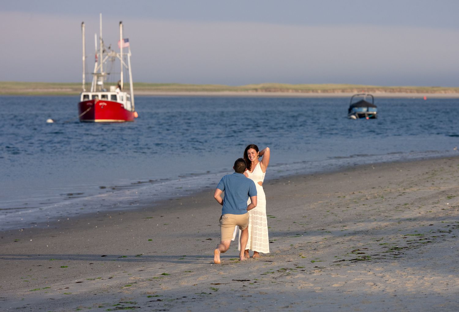 Couple embracing on a sandy beach at sunset with a red fishing boat anchored in calm waters behind them.