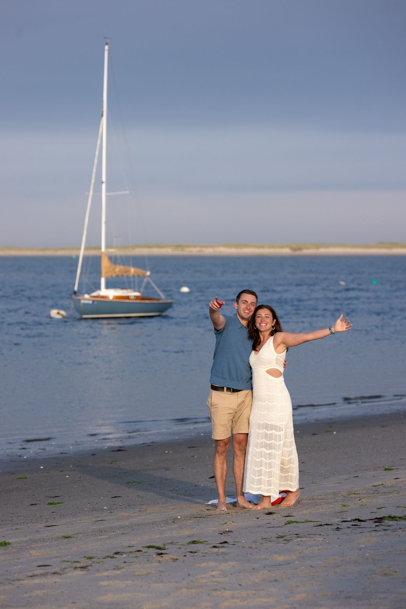 Happy couple giving thumbs up on sandy beach at sunset with sailboat anchored in calm blue water behind them.