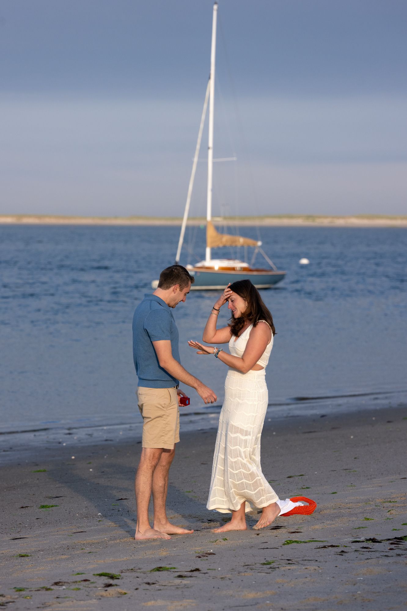 Couple on a sandy beach at sunset with a sailboat in the background, man presenting a ring box to surprised woman.
