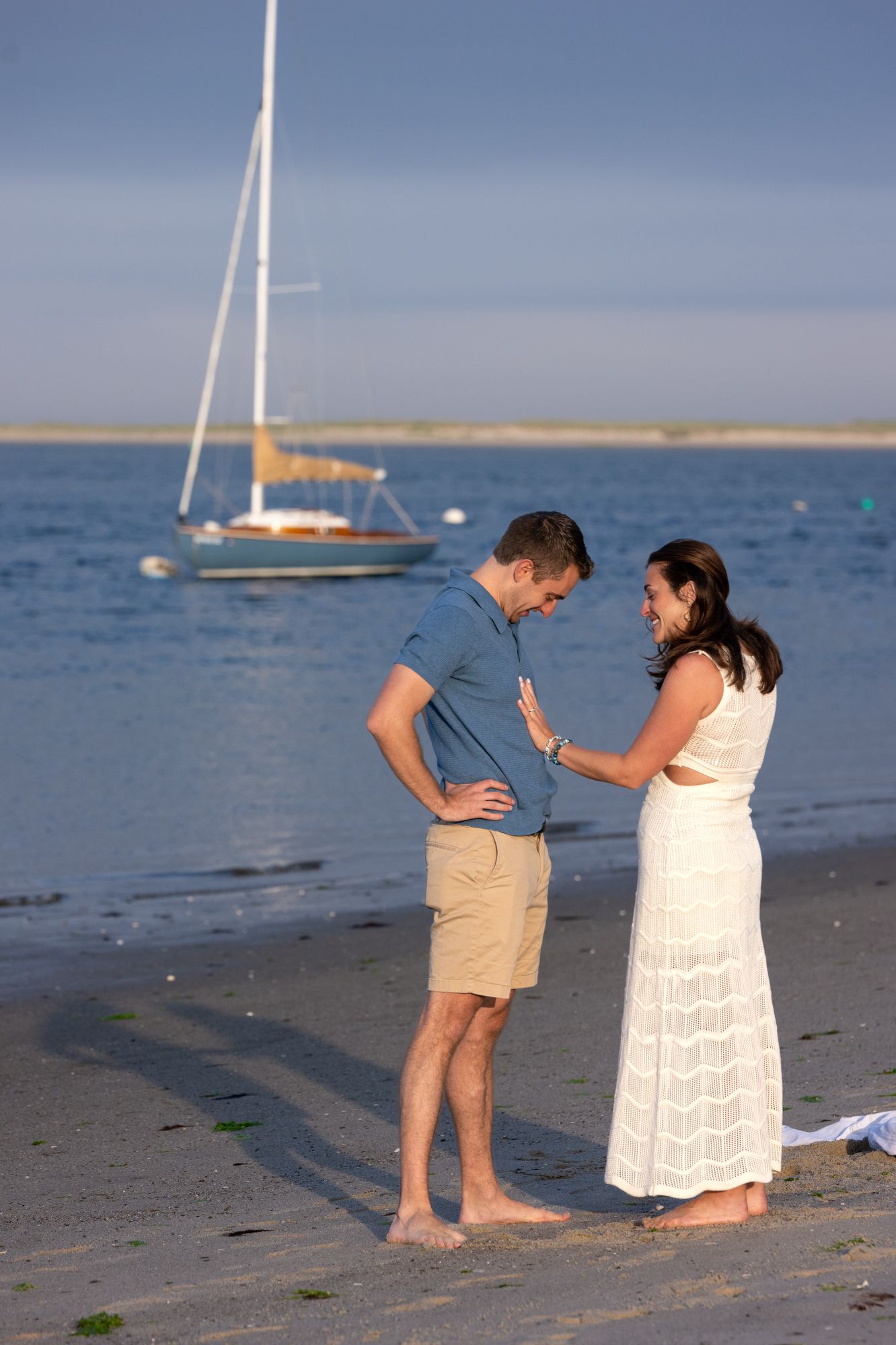 Couple sharing a romantic moment on a sandy beach at sunset with a sailboat anchored in the calm blue water behind them.