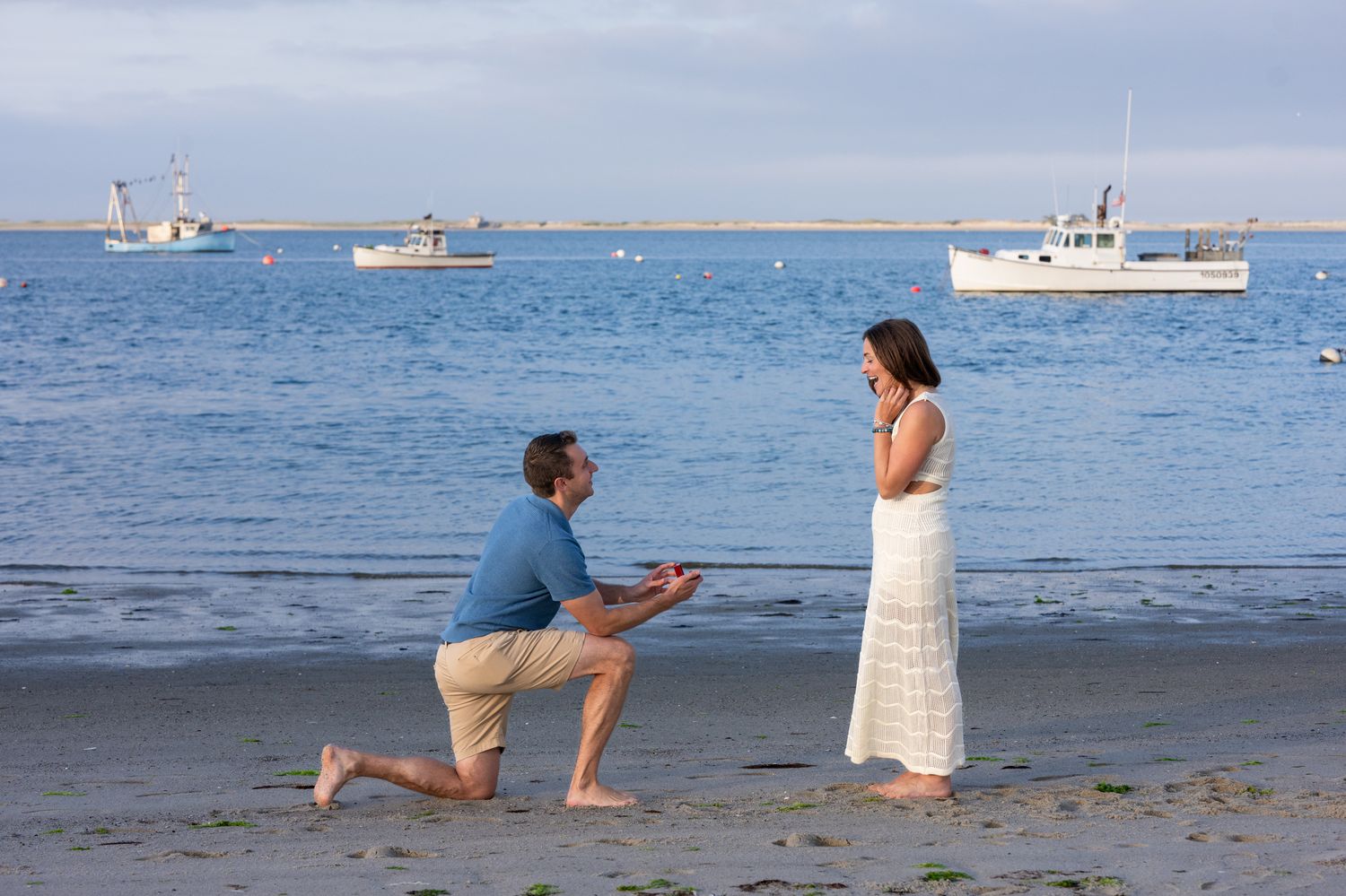 Man proposing to woman on a sandy beach with boats in the background on a calm blue ocean.