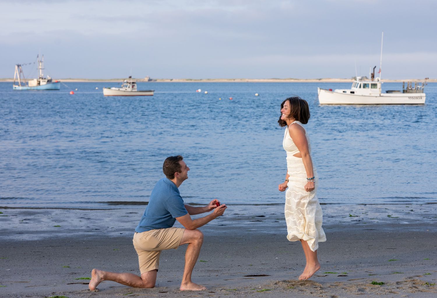 Man proposing on bended knee to woman on sandy beach with boats in calm harbor in background.