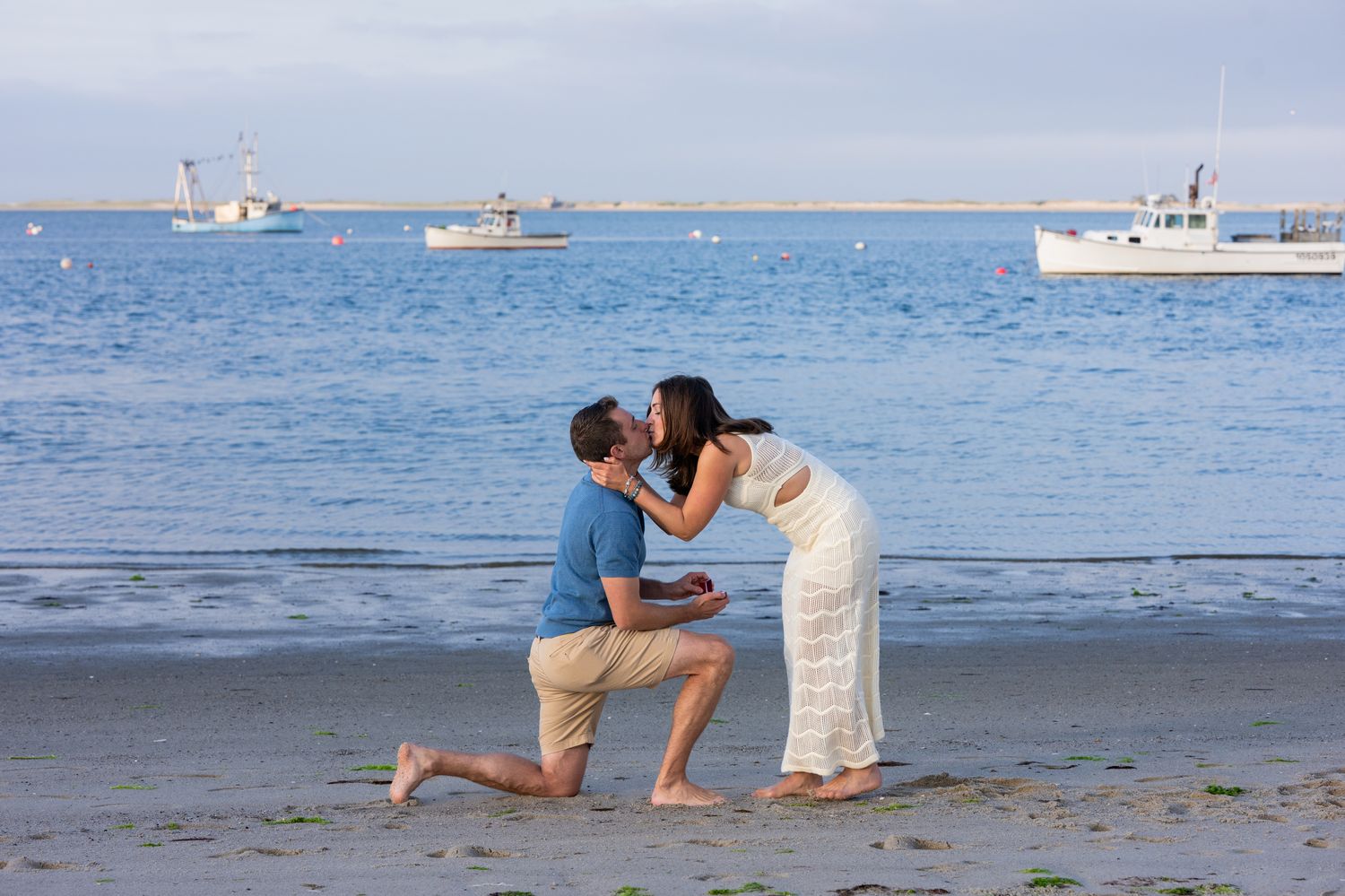 Man kneeling on beach proposing to woman in white dress, with boats on calm ocean waters in background.