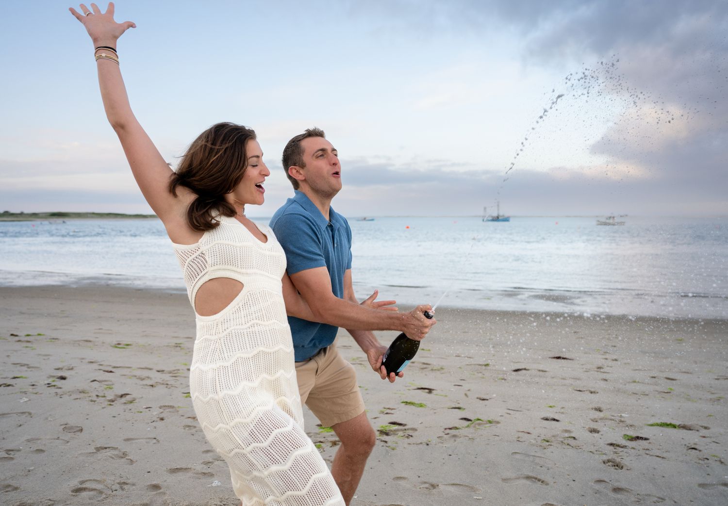 Joyful couple celebrating on a beach, woman in white dress with arms raised as man pops champagne bottle.