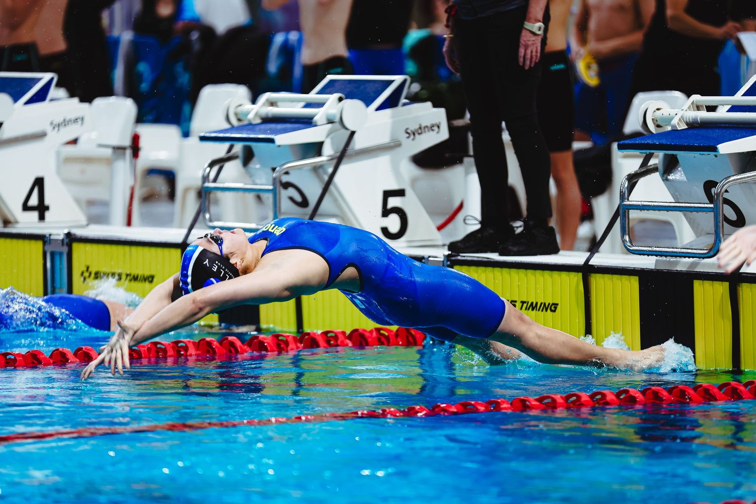 Female swimmer in purple swimsuit diving into pool at competitive swimming event with lane markers visible.