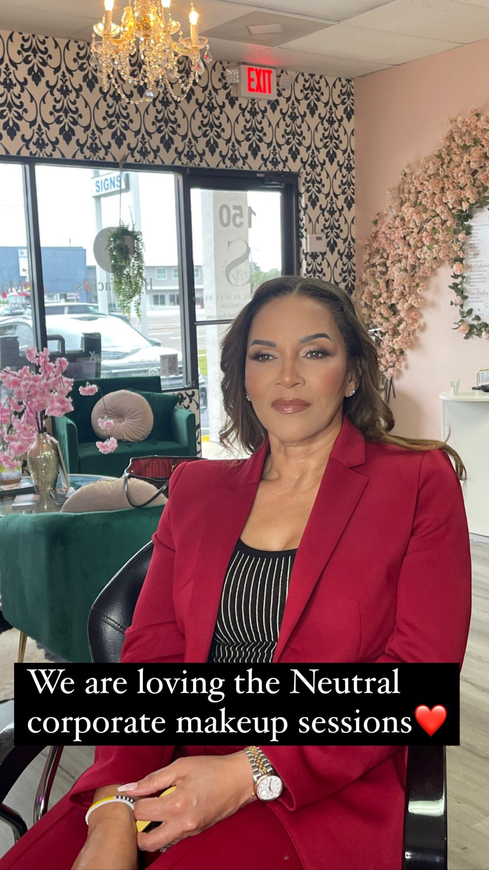 Woman in red blazer sits in salon chair showcasing neutral corporate makeup look in decorated beauty studio.