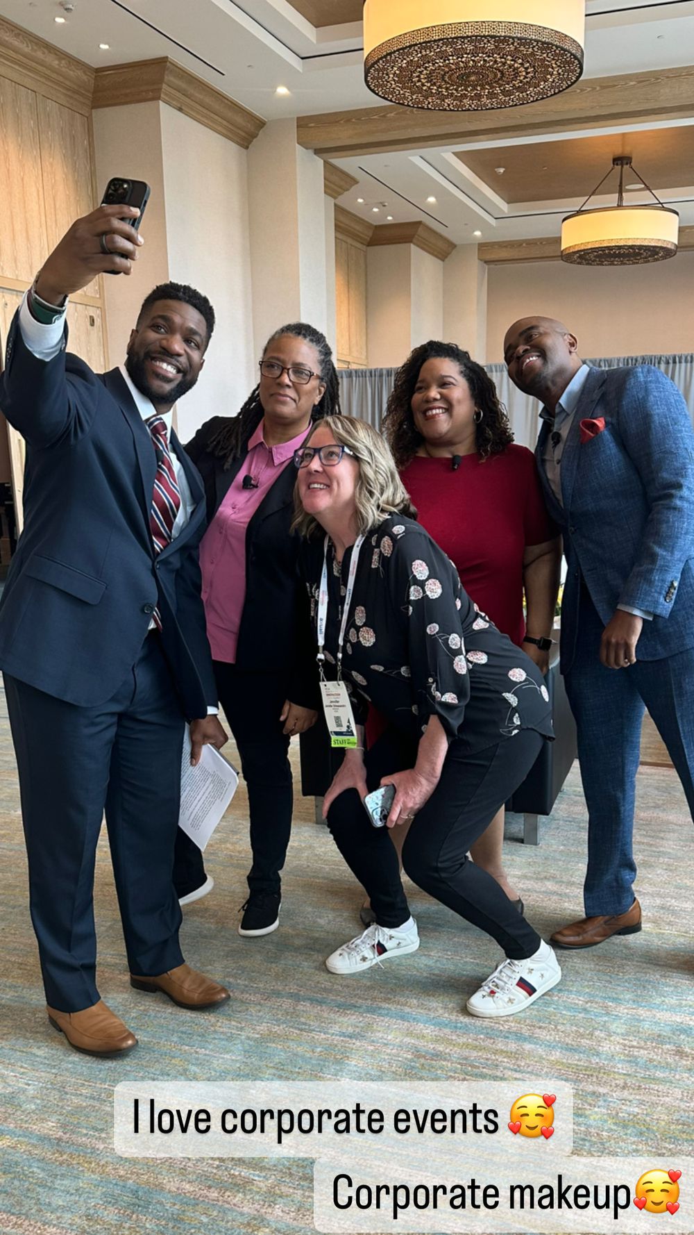 Five professionals pose for a selfie at a corporate event in an elegant hotel ballroom, smiling and dressed in business attire.
