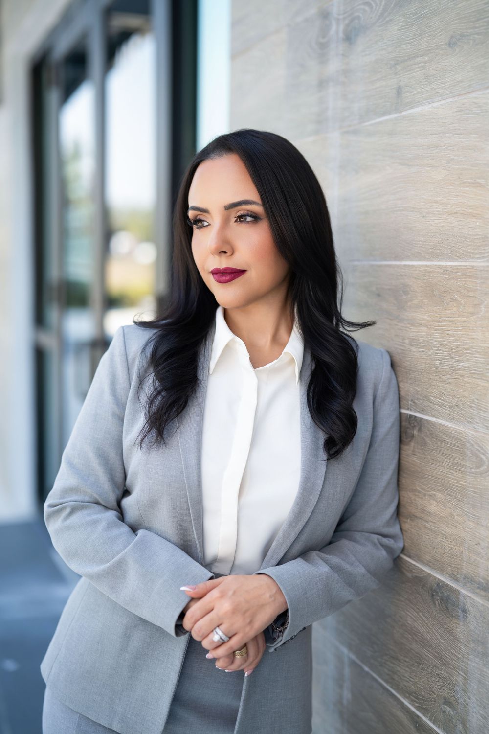 Professional woman in gray blazer and white blouse leaning against a wall outdoors.