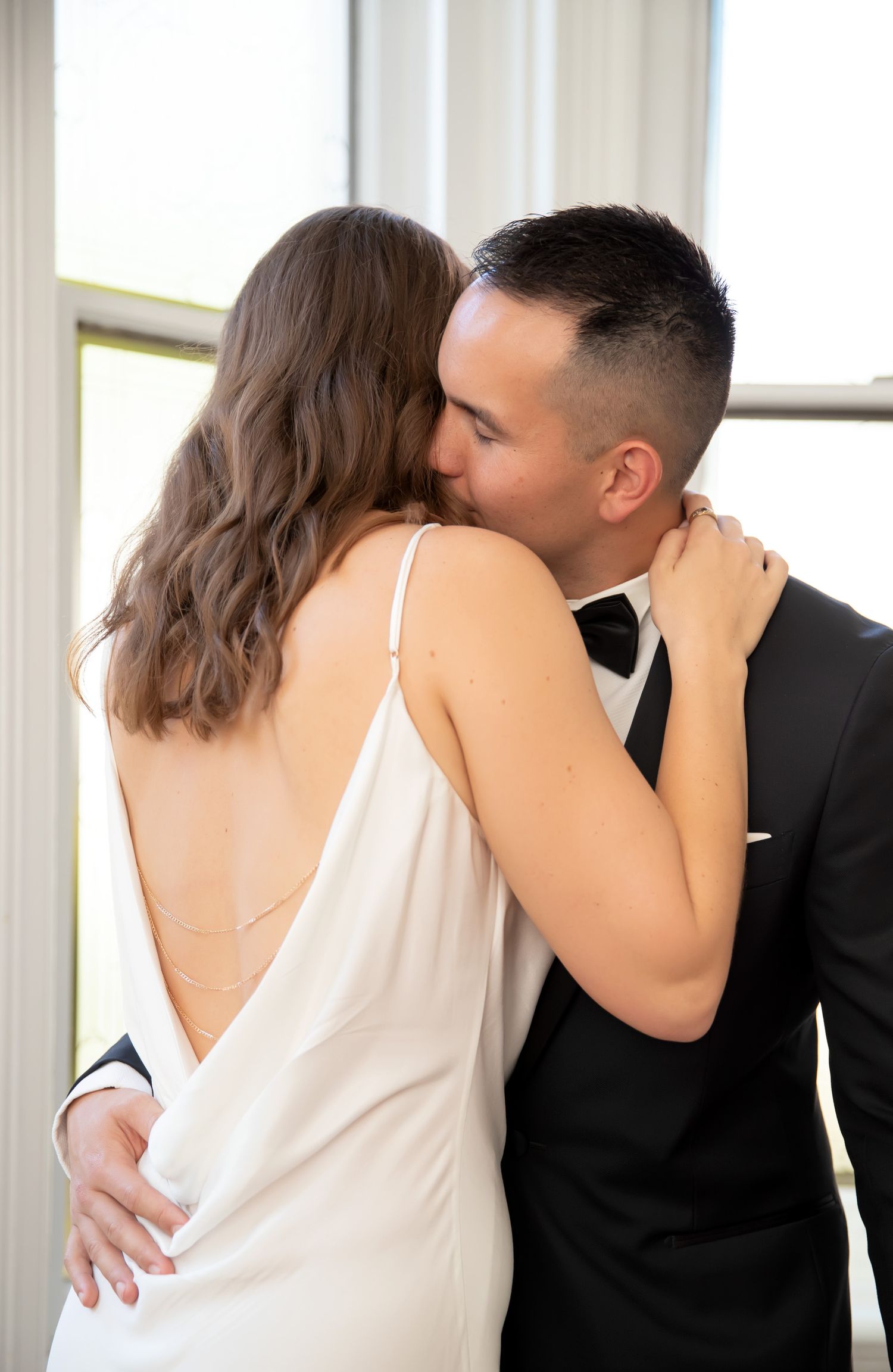 A bride in a backless white slip dress and groom in a black tuxedo share a romantic kiss near a bright window.