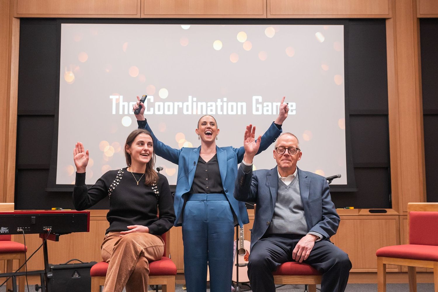 Three people raise their hands joyfully on stage in front of a screen at a Professor Alison Wood Brooks Harvard Business School event.