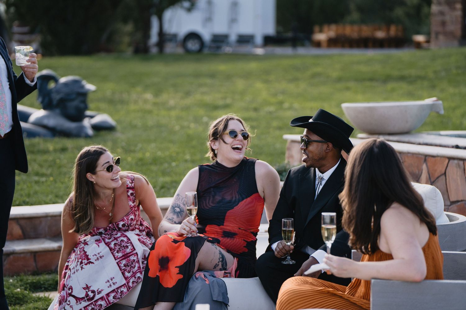 Four elegantly dressed guests socializing outdoors at a garden party, enjoying champagne on a modern stone bench.