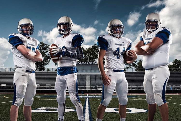 Four high school football players posing in white and blue uniforms on a stadium field.