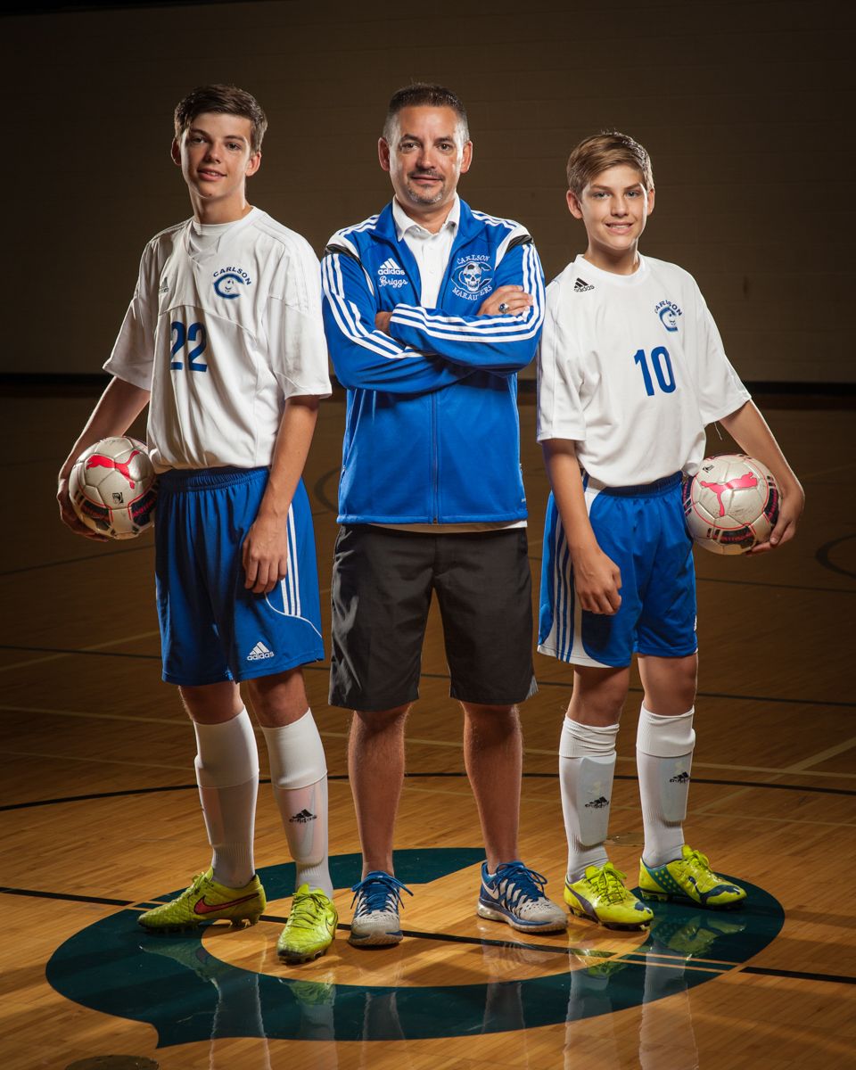 Youth soccer coach stands between two players holding balls in gymnasium team portrait.