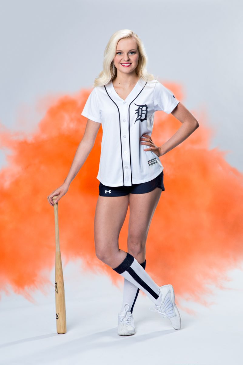 Blonde athlete poses with baseball bat in Detroit Tigers white jersey against orange smoke background.