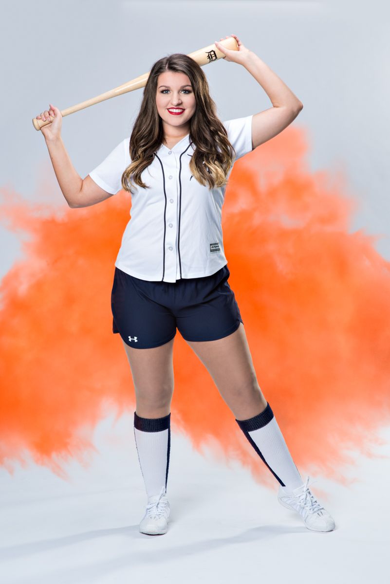 Young woman in Detroit Tigers baseball uniform holds bat behind shoulders in orange smoke backdrop.