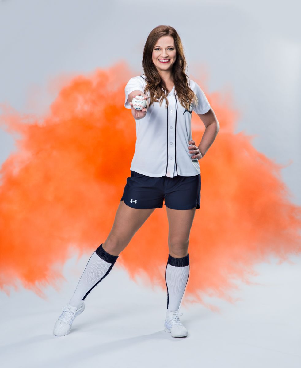 Female softball player in uniform holding a ball with orange smoke.