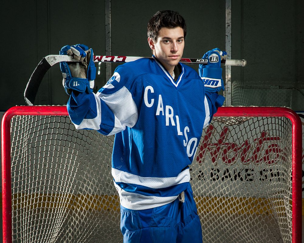 Male hockey player in blue Carlson jersey poses with stick at goal net in dramatic portrait.