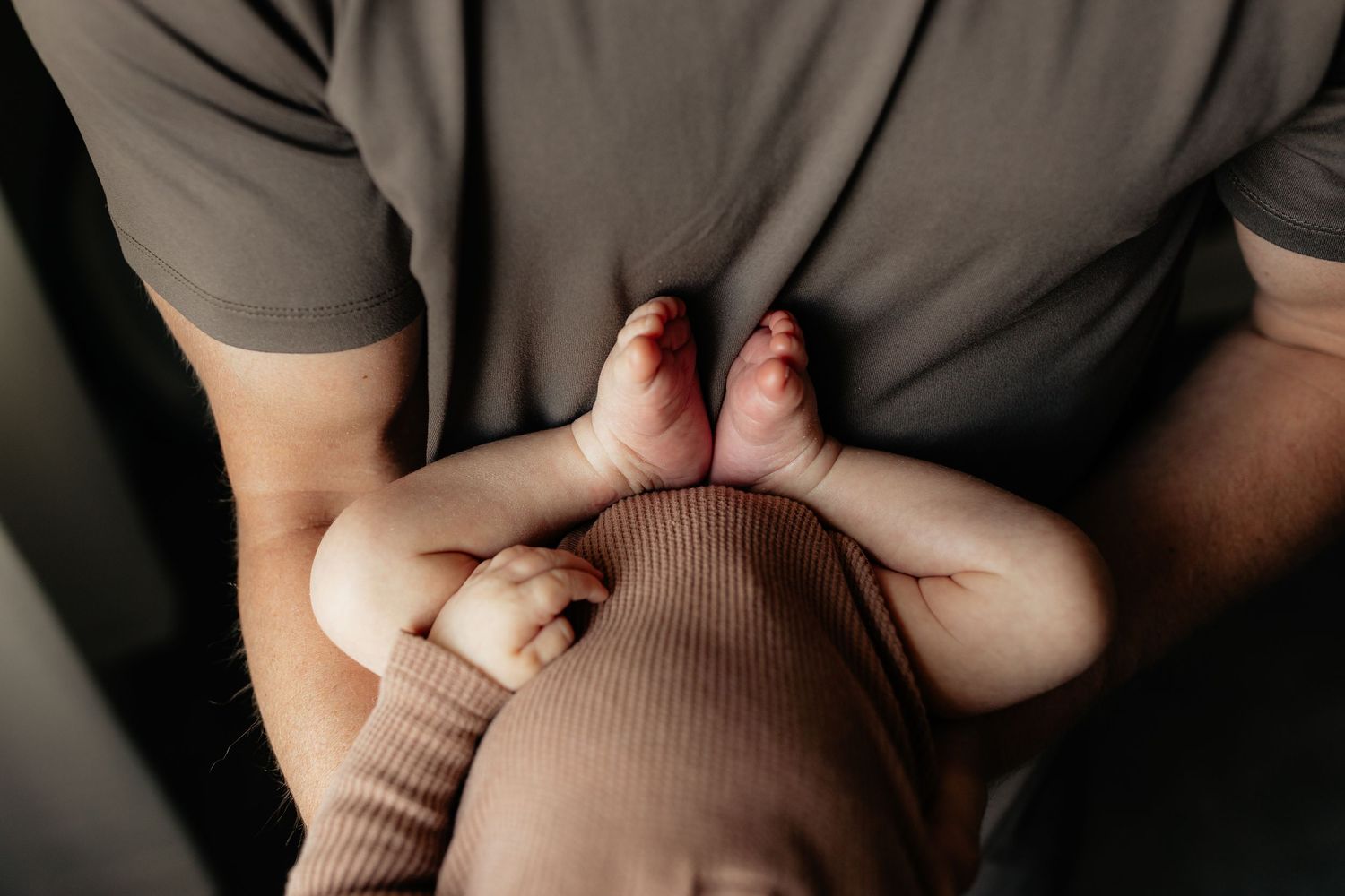 Close-up of a parent in gray shirt cradling a newborn baby's tiny feet and hands on their chest.
