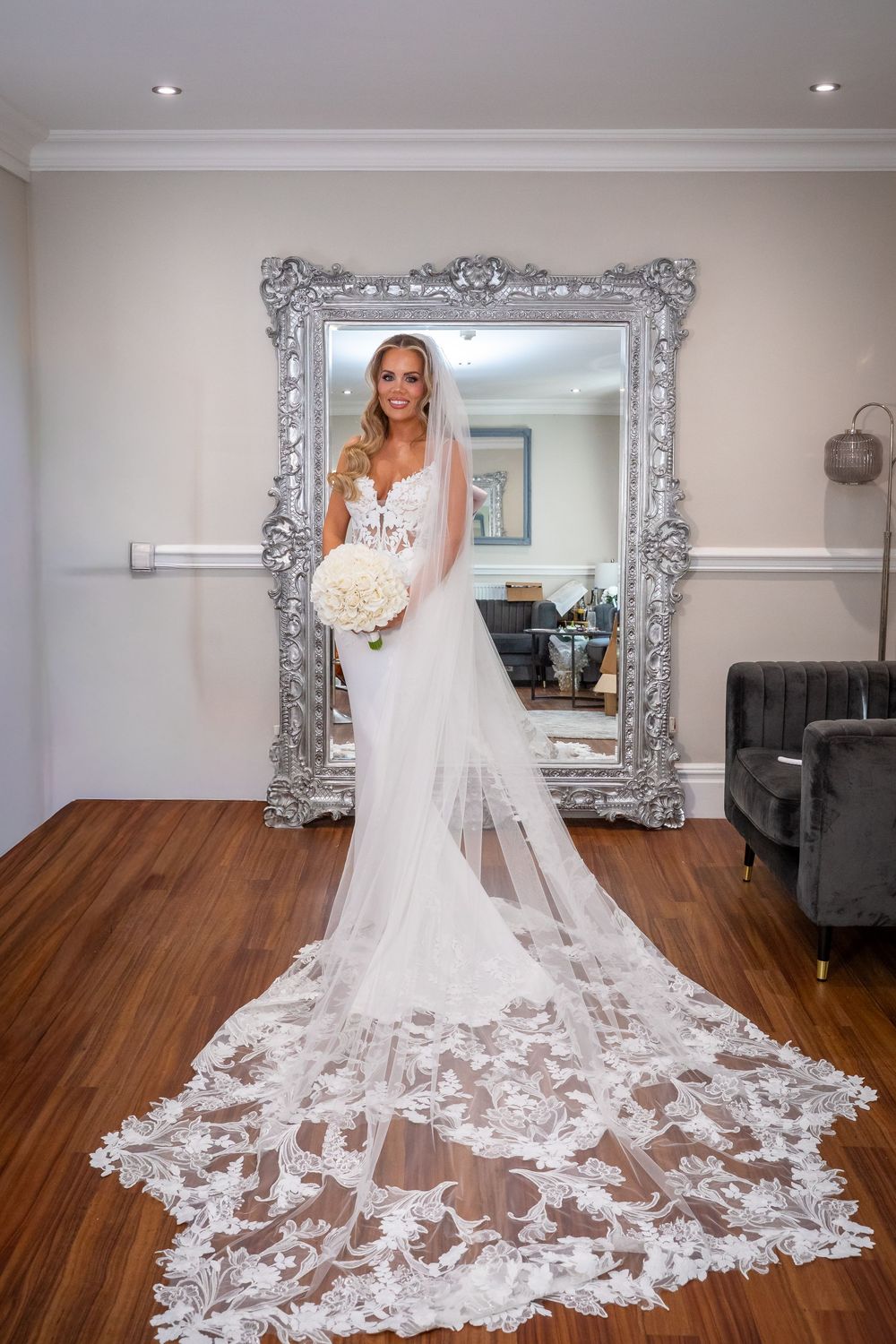 Bride in white lace wedding gown with cathedral veil holding bouquet, standing before ornate silver mirror on hardwood floor.