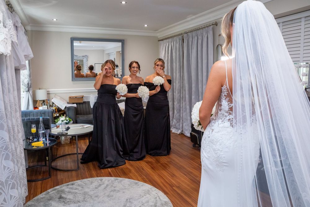 Bride in lace wedding gown and veil stands before three bridesmaids in black dresses reacting with joy in a hotel room.