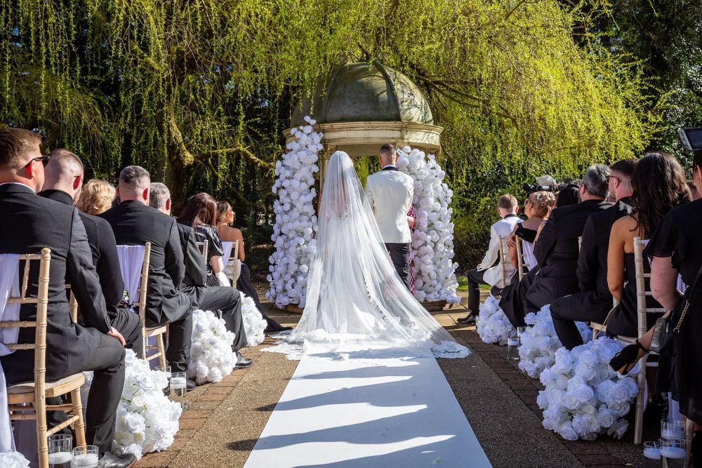 Elegant outdoor wedding ceremony with bride walking down white aisle toward floral arch gazebo, guests seated on both sides.