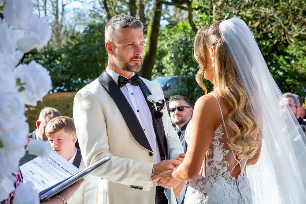 A groom in a white tuxedo exchanges vows with a bride in a lace gown during an outdoor wedding ceremony.