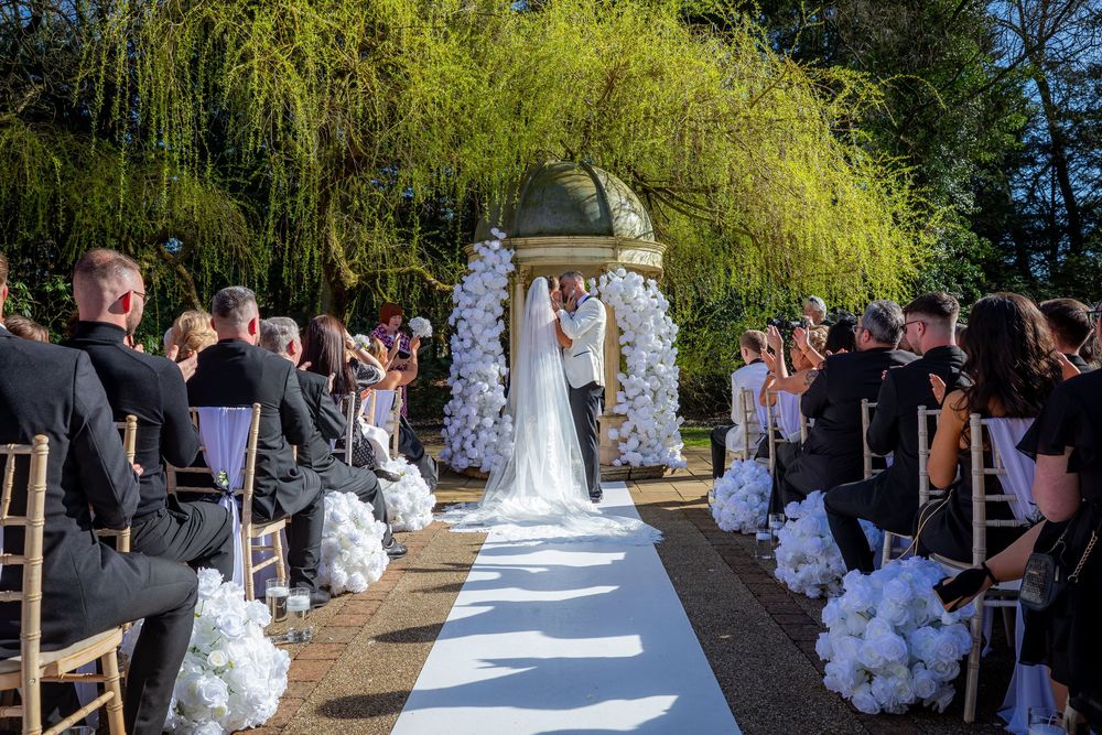 Elegant outdoor wedding ceremony with white floral arch, aisle runner, and guests seated on wooden chairs under a willow tree.