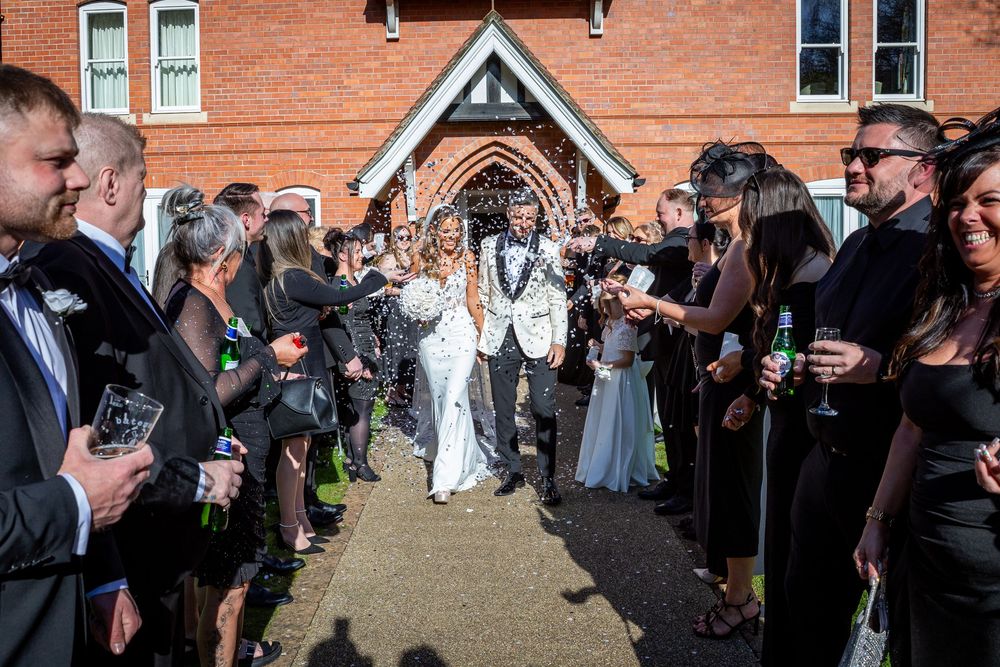 Bride and groom walk through confetti tunnel with smiling guests outside a red brick church on their wedding day.