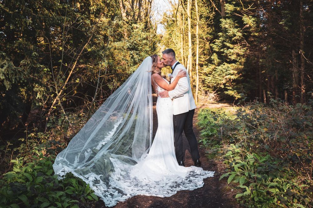 Bride in fitted gown with lace veil and groom share a kiss in a lush forest setting on their wedding day.