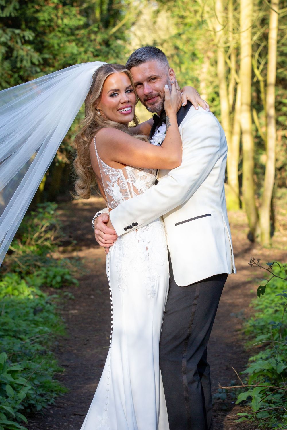 Bride and groom embracing on a woodland path, bride in lace gown with veil, groom in white tuxedo jacket.