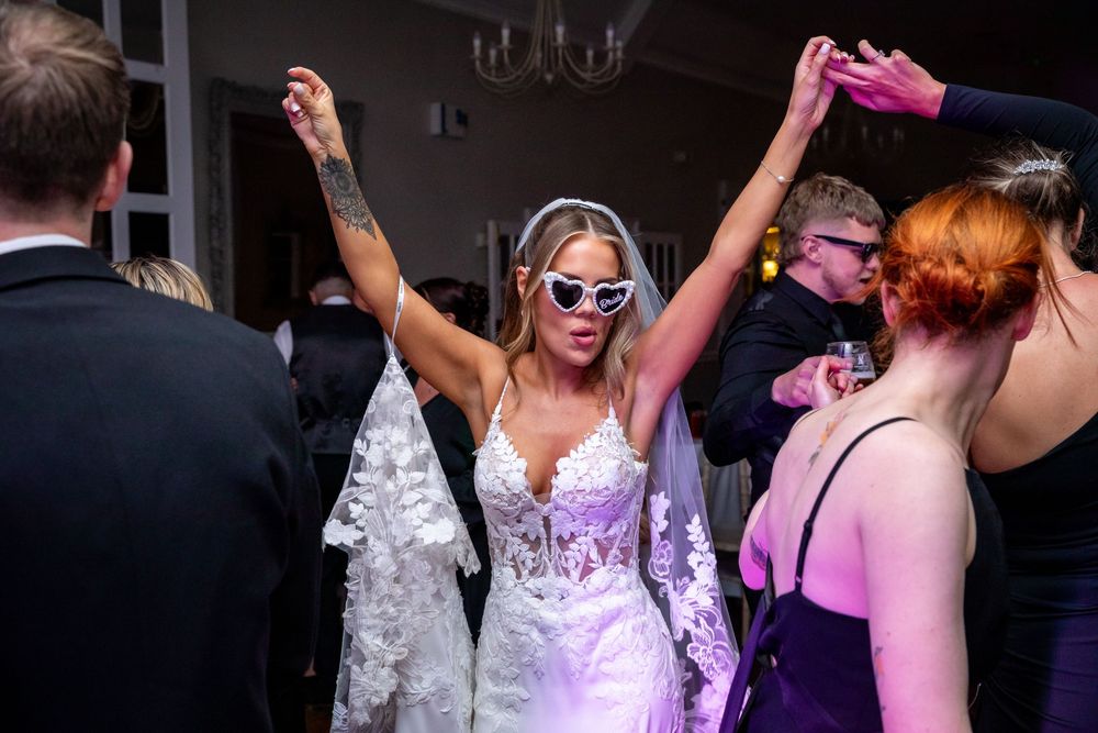 Bride in lace wedding dress and heart sunglasses dancing joyfully with arms raised at wedding reception.