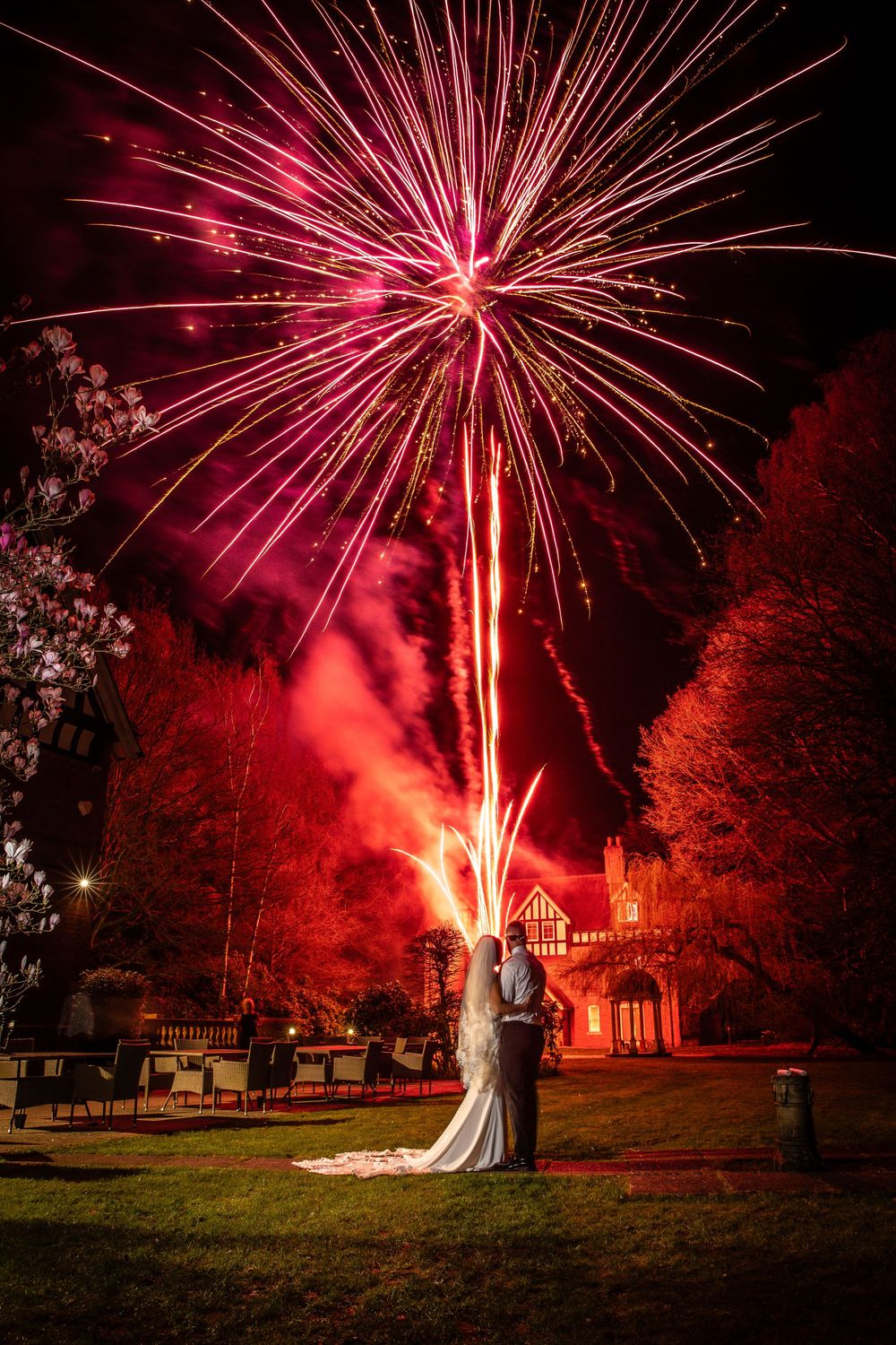 Bride and groom embrace as vibrant red fireworks explode over a Tudor mansion during their romantic outdoor wedding celebration.