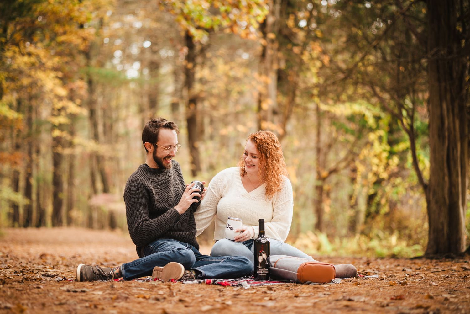 Engaged couple laughing together on a blanket in the forest at Borderland State Park, toasting with personalized 'Mr.' and 'Mrs.' mugs