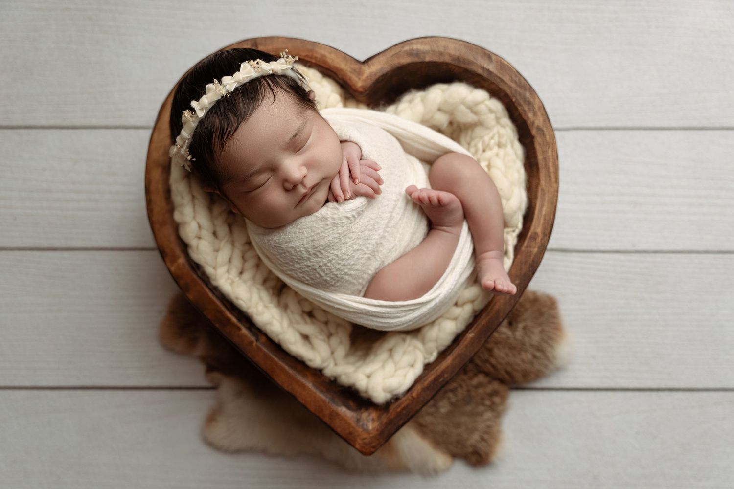 Newborn baby girl sleeping peacefully in a heart-shaped wooden bowl with cream knit wrap and floral headband.