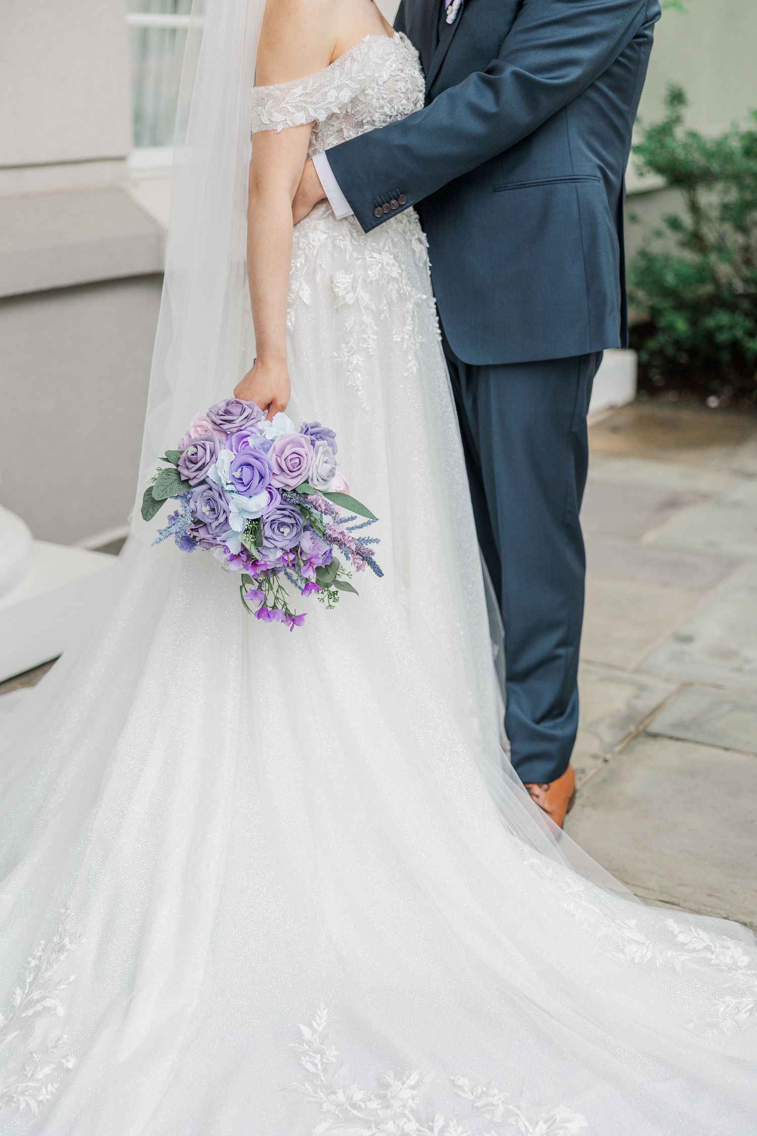 Bride in lace gown holding purple rose bouquet embraces groom in navy suit on wedding day.